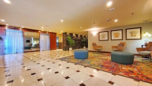 Un hall d’entrée spacieux avec un sol à motifs, un grand pouf bleu, des fauteuils en osier et un mur en pierre agrémenté de plantes.