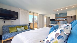 Quarto de hotel moderno com uma cama grande, um sofá, uma TV, varanda com vista para a cidade e uma cozinha compacta.