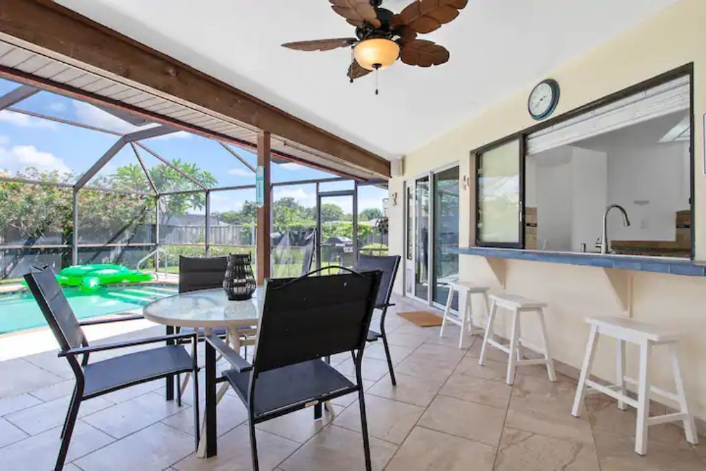 A covered patio area with a glass enclosure, a round table with chairs, a ceiling fan, and a view of a swimming pool and greenery.