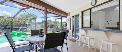 A covered patio area with a glass enclosure, a round table with chairs, a ceiling fan, and a view of a swimming pool and greenery.