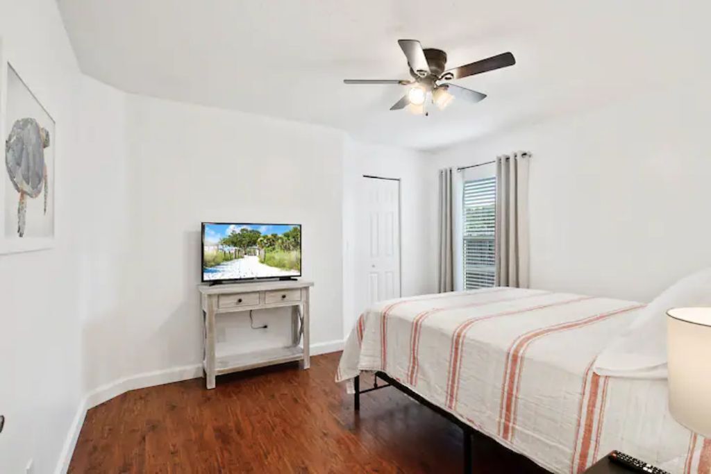 A bedroom with a bed, a television on a small white cabinet, a ceiling fan, and a window with blinds.