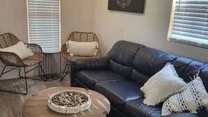 A living room with a black leather sofa, two wicker chairs, a round coffee table with a fire pit, and white blinds on the windows.