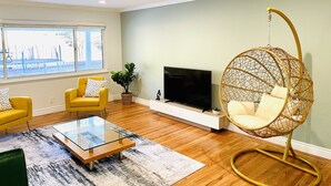 Una sala de estar con un sofá amarillo, una mesa de centro de cristal, una silla colgante, un televisor y un ventanal grande.