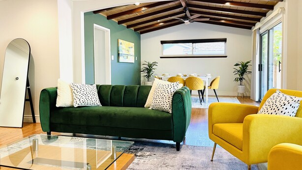 Una sala moderna con un sofá verde, sillones amarillos, una mesa de centro de vidrio y un área de comedor al fondo.