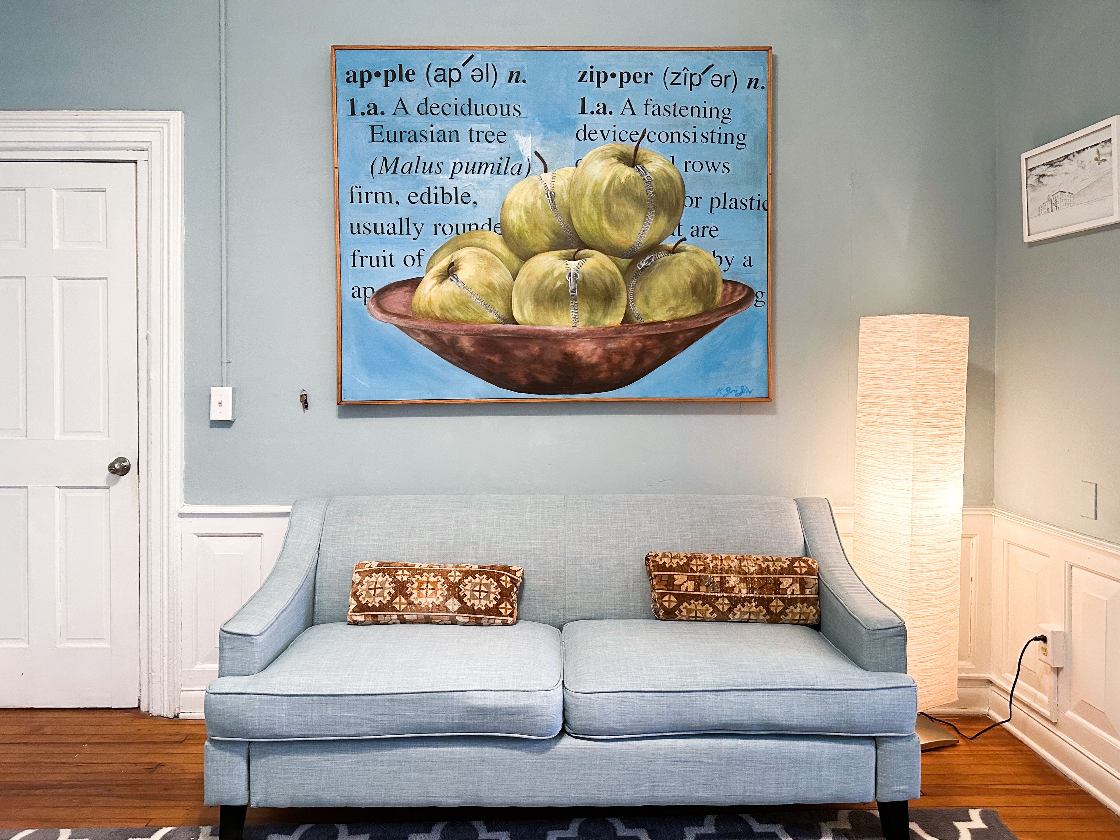 Un salón con un sofá azul claro, dos cojines estampados, un cuadro de manzanas en la pared y una lámpara.