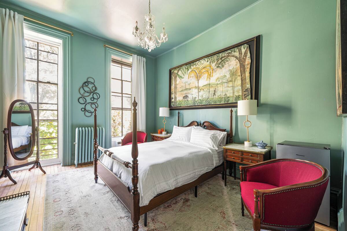 A bedroom with a four-poster bed, a red armchair, a dresser, a nightstand, a desk, a radiator, a chandelier, and a large landscape painting on the wall.
