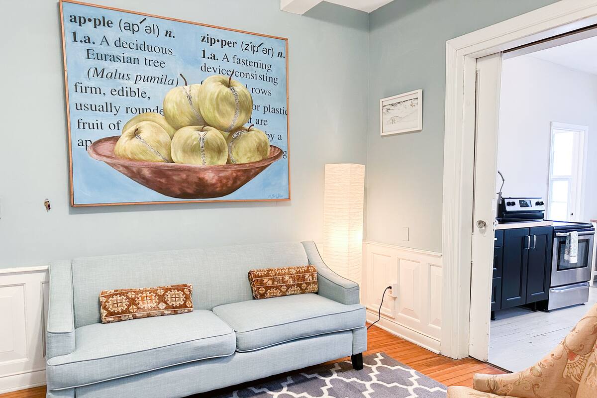 A living room with a light blue sofa, patterned cushions, a rug, and a wall with a large apple illustration.