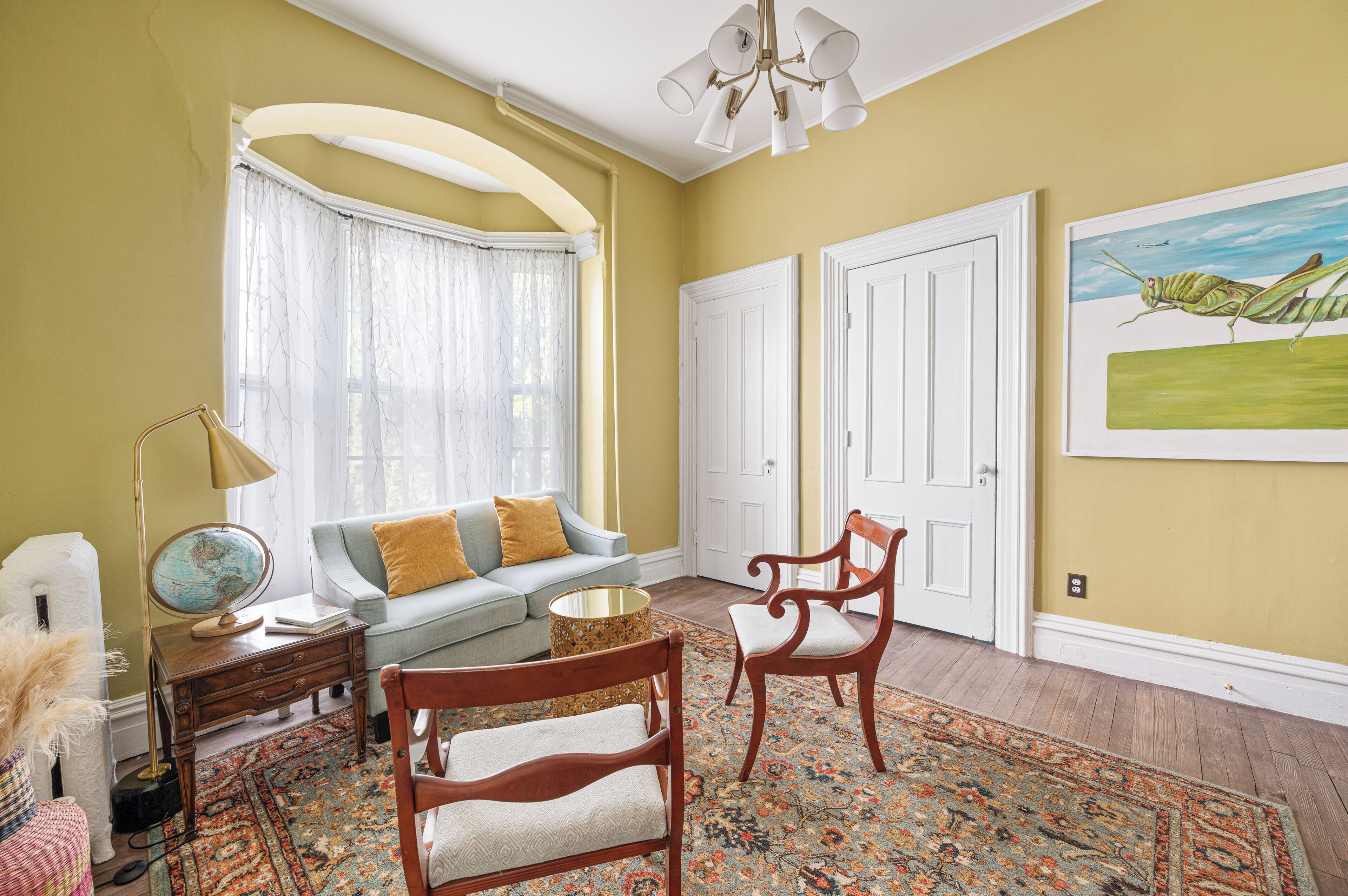 A room with a light yellow wall, a globe on a side table, a sofa with cushions, a wooden chair, a patterned rug, and a painting of a grasshopper.