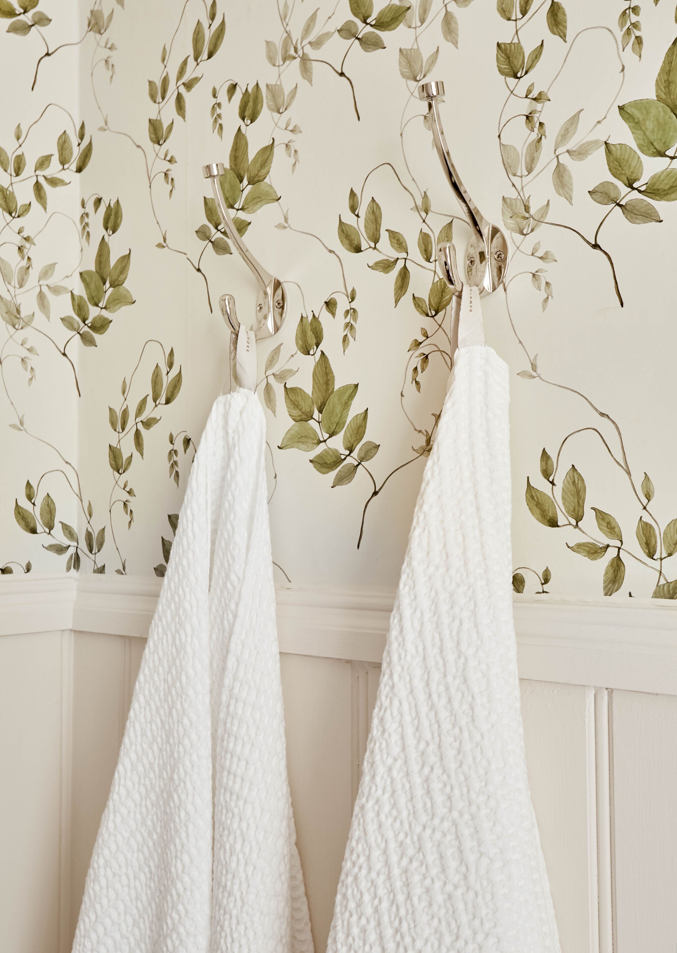 Une salle de bain avec une serviette blanche à texture particulière suspendue à un porte-serviettes, le tout adossé à un mur recouvert d’un papier peint à motifs de feuilles.