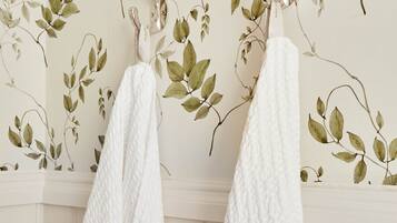 Une salle de bain avec une serviette blanche à texture particulière suspendue à un porte-serviettes, le tout adossé à un mur recouvert d’un papier peint à motifs de feuilles.