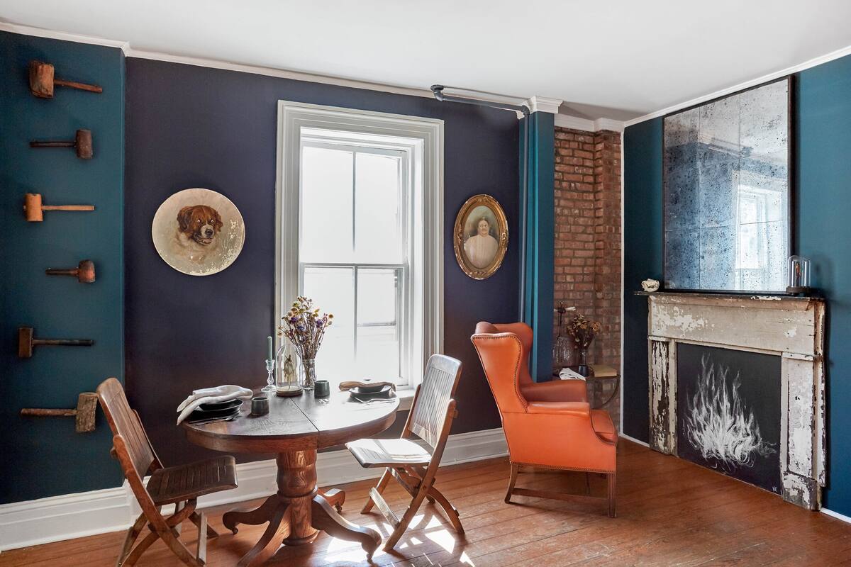 A dining room with a round table, two wooden chairs, a decorative hat on the wall, and a fireplace with a painted design.