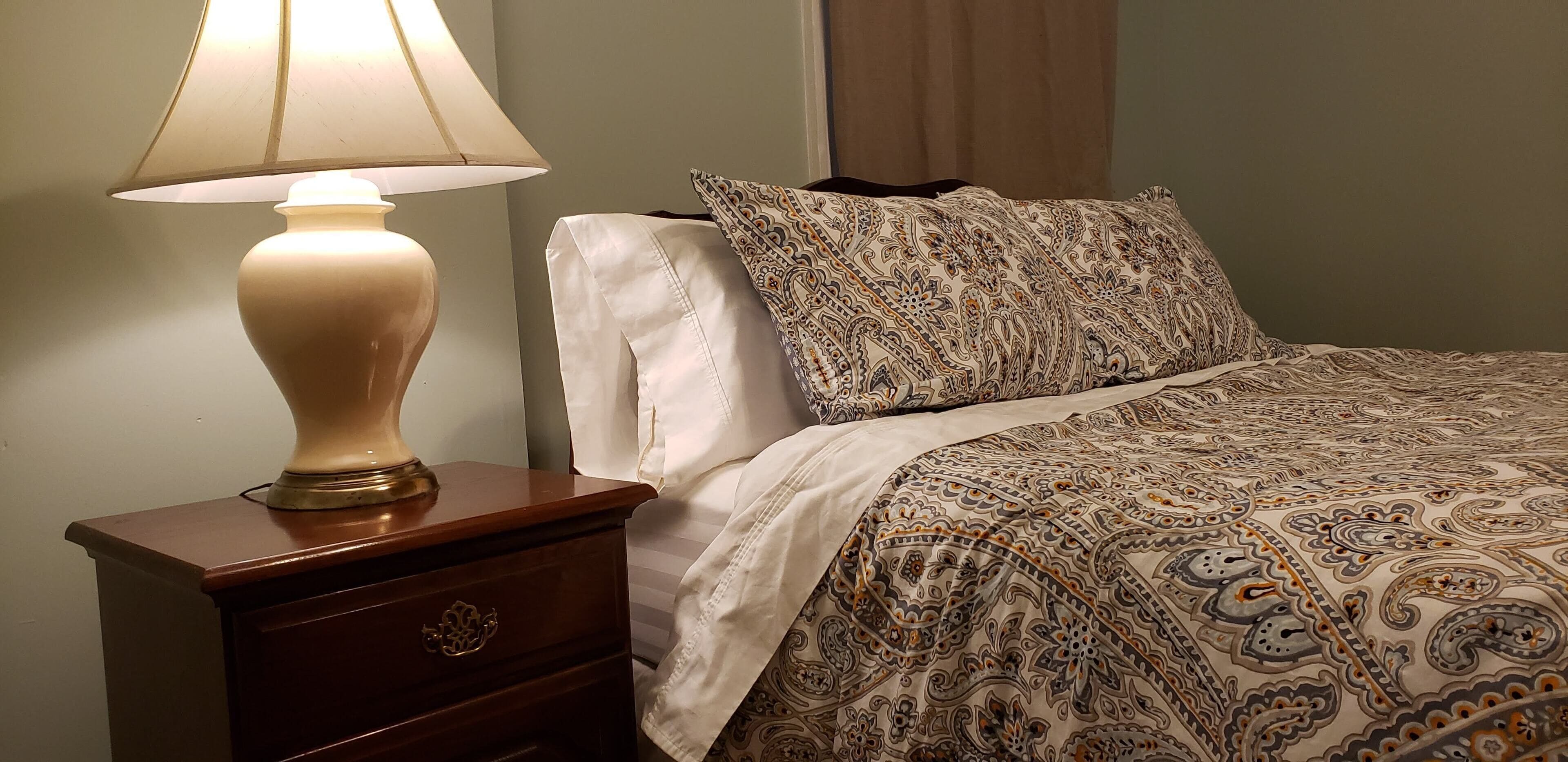 Een netjes opgemaakt bed met een geborduurde spreideken, een bedlampje en een houten nachtkastje met een lade.
