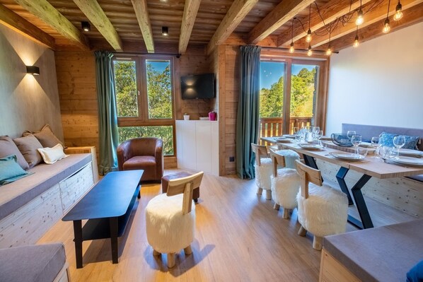 A cozy cabin-style room with a wooden ceiling, a sofa, a dining table set for four, and a large window with curtains.