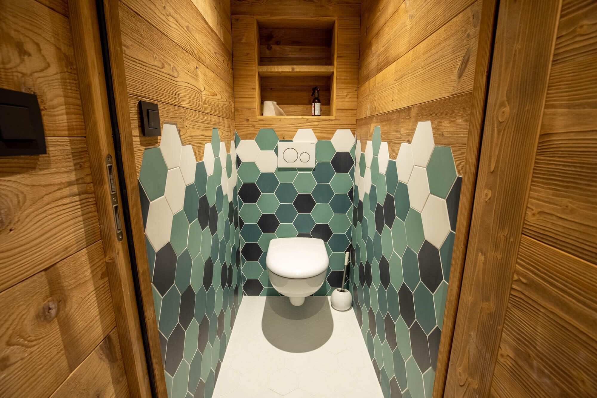 Een kleine badkamer met houten lambrisering, een wit toilet, hexagonale tegels en een plankje met een fles en een klein voorwerp.
