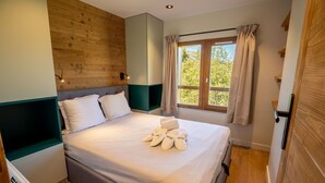 A neatly made bed with white linens and towels, a wooden headboard, a window with curtains, and a view of greenery outside. - Hauts de Chantemouche 17 (Les Allues)