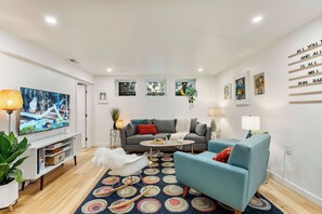 Un salon moderne avec un canapé gris, un fauteuil bleu, une table basse ronde et un téléviseur placé à gauche. On y trouve des plantes, des œuvres d’art accrochées aux murs et un tapis à motifs sur le sol.