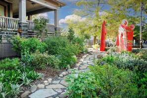 Un chemin de jardin menant à une maison dotée d’un porche, entourée de différentes plantes et agrémentée d’une sculpture rouge.