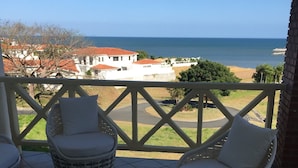 Ein Balkon mit Korbstühlen und einem runden Tisch, mit Blick auf die Küste, wo Gebäude und eine Straße zu sehen sind.