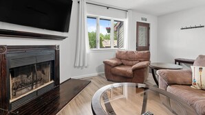 A living room with a fireplace, a television, a brown leather armchair, a glass coffee table, and a view of the outdoors through a window.