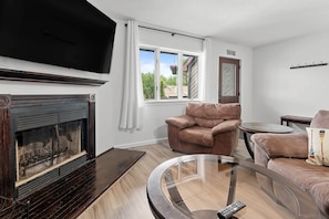 A living room with a fireplace, a television, a brown leather armchair, a glass coffee table, and a view of the outdoors through a window.