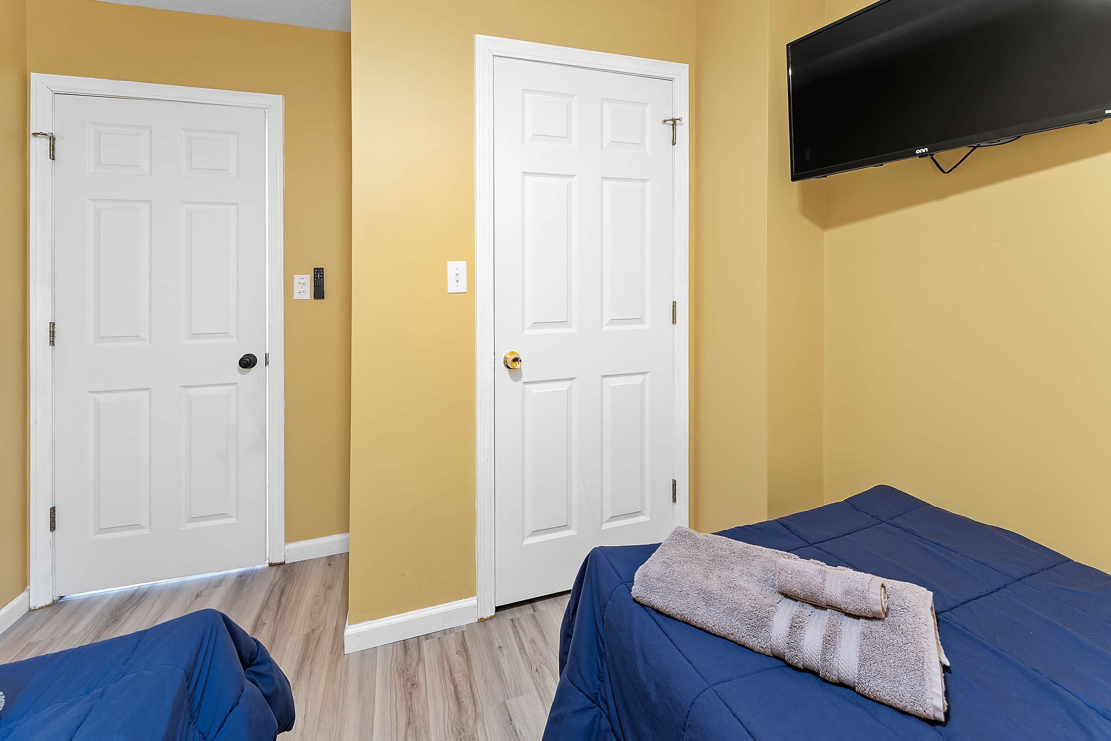 Una habitación con dos puertas blancas, una cama con colcha azul, un televisor montado y un interruptor de luz en la pared.