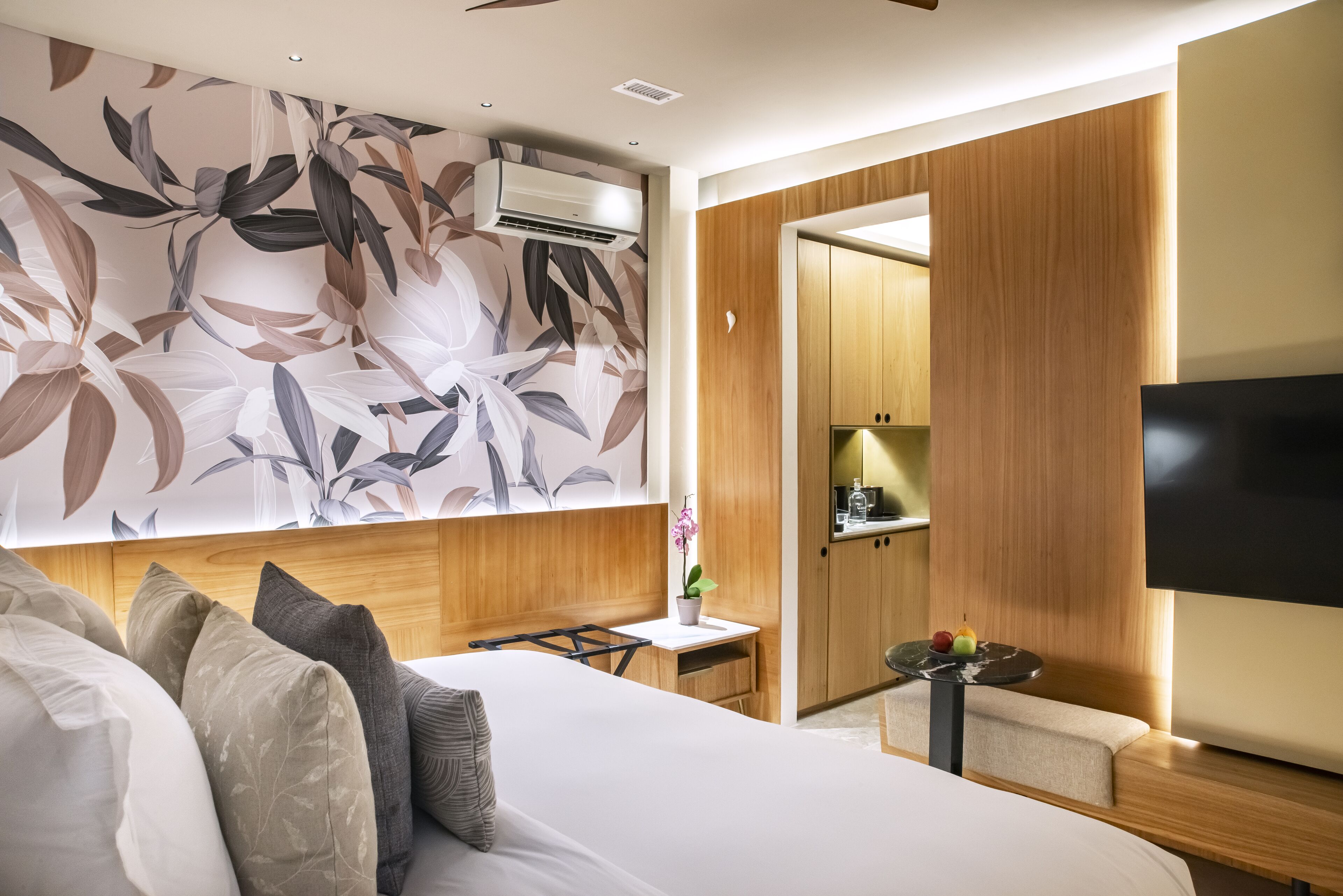 A modern hotel room with a large bed, a wooden headboard, a flat-screen TV, a small table with fruit, and a decorative wall with a floral pattern.