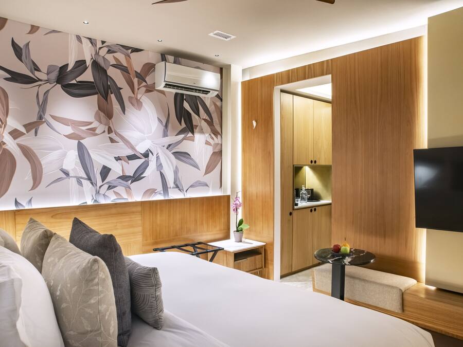 A modern hotel room with a large bed, a wooden headboard, a flat-screen TV, a small table with fruit, and a decorative wall with a floral pattern.