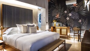 A modern hotel room with a large bed, a wooden headboard, a wall-mounted air conditioner, a small desk with a chair, and a decorative plant.