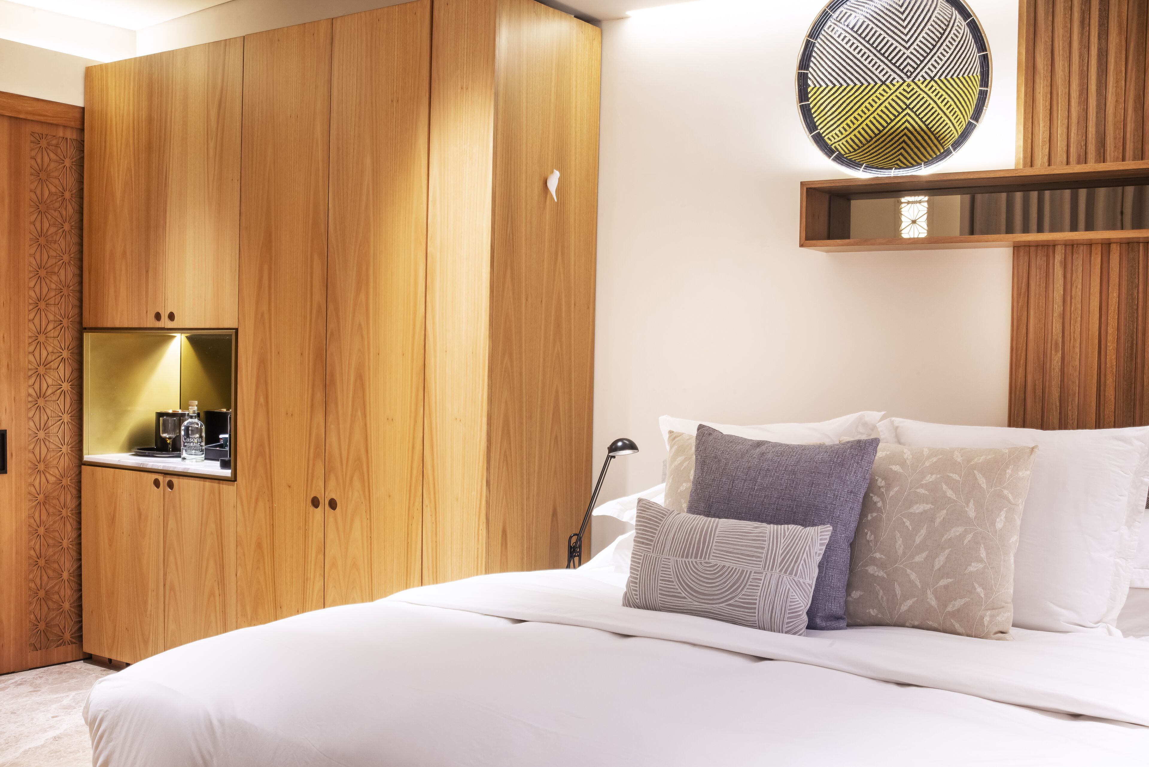 Un dormitorio con un armario de madera, una cama con varias almohadas, una mesita de noche con lámpara y un espejo de pared.