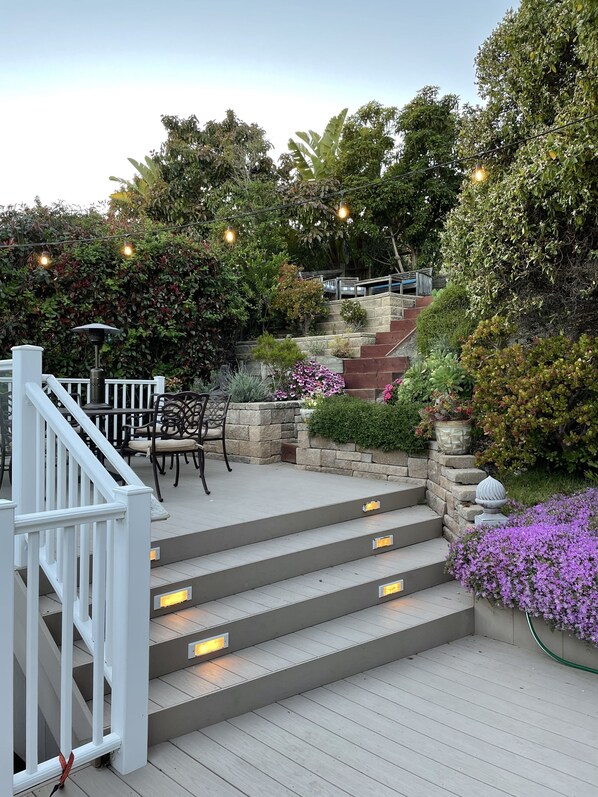 Une terrasse en bois avec des marches menant à un espace détente, entourée d’une végétation luxuriante et de plantes en fleurs.