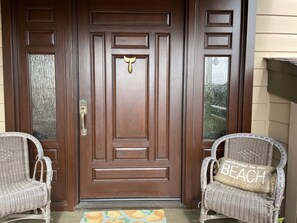 A wooden front door with a brass handle, flanked by two wicker chairs with cushions.