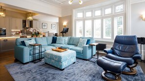 A modern living room with a sectional sofa, a leather armchair, and a coffee table. There is a kitchen in the background with a stove and cabinets. - Tranquil Apartment in Merseyside (Liverpool)
