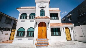 Un edifici de color blanc amb una entrada central, un cartell sobre la porta i un teulat de rajoles.