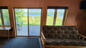 Una habitación con una mesa de madera, un sofá estampado y amplios ventanales que ofrecen vista a un lago y vegetación.