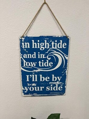 A blue sign with white text saying "in high tide and in low tide I'll be by your side" on a white wall.