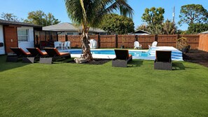 A backyard with artificial grass, a pool, lounge chairs, and a wooden fence.