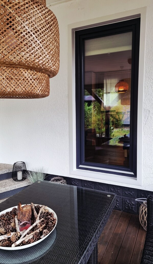 A modern outdoor seating area with a glass table, wicker hanging light fixture, and a view of a garden through a large window.