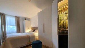 Una camera d'albergo moderna con un letto, un poggiapiedi blu, un armadio e un quadro raffigurante una scena boschiva.