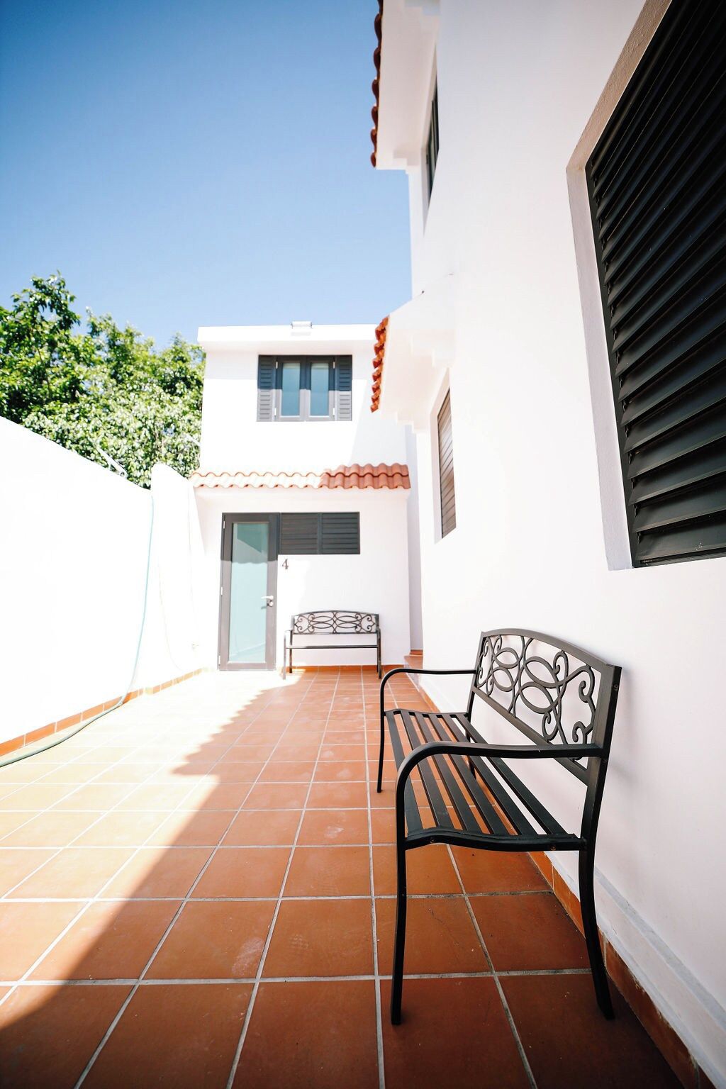 Un patio con un banco de metal negro y una mesita, paredes blancas y piso de baldosas.