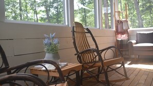 Une chaise à bascule en bois et une table sur laquelle est posée une plante en pot, le tout sur une terrasse en bois dotée de grandes fenêtres.