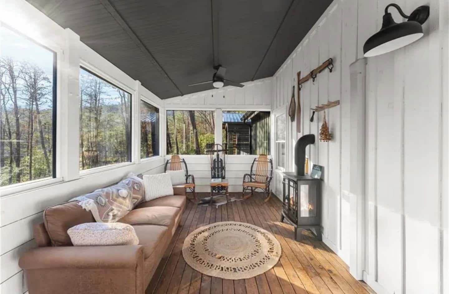 Eine überdachte Veranda mit Holzboden, einer Couch, zwei Schaukelstühlen, einem Tisch und einem Holzofen.