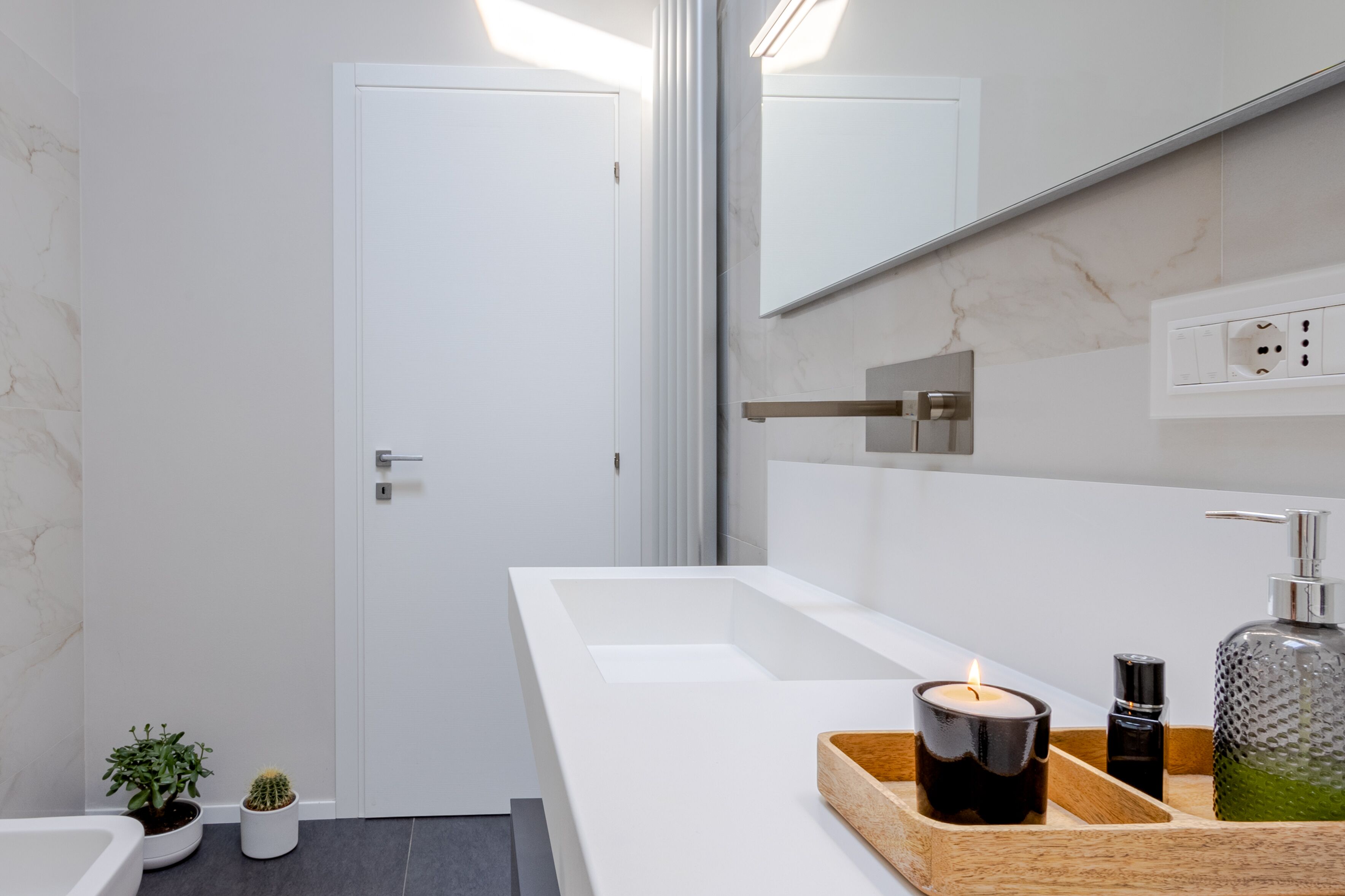 Un bagno moderno con un lavandino bianco, uno specchio e un vassoio di legno con candele e articoli da toilette.