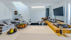 Un salon moderne avec un canapé, une table basse sur laquelle sont disposés des fruits, un coin cuisine avec un téléviseur et un escalier.