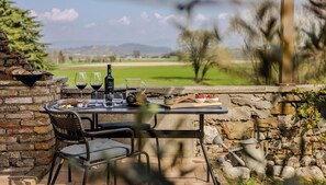 Zona pranzo all'aperto con parete in pietra, tavolo apparecchiato con bicchieri da vino, una bottiglia e cibo.