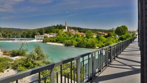 Un pont doté d’une rambarde métallique surplombant une rivière, entouré de verdure et de bâtiments, dont un se distingue par sa haute tour.