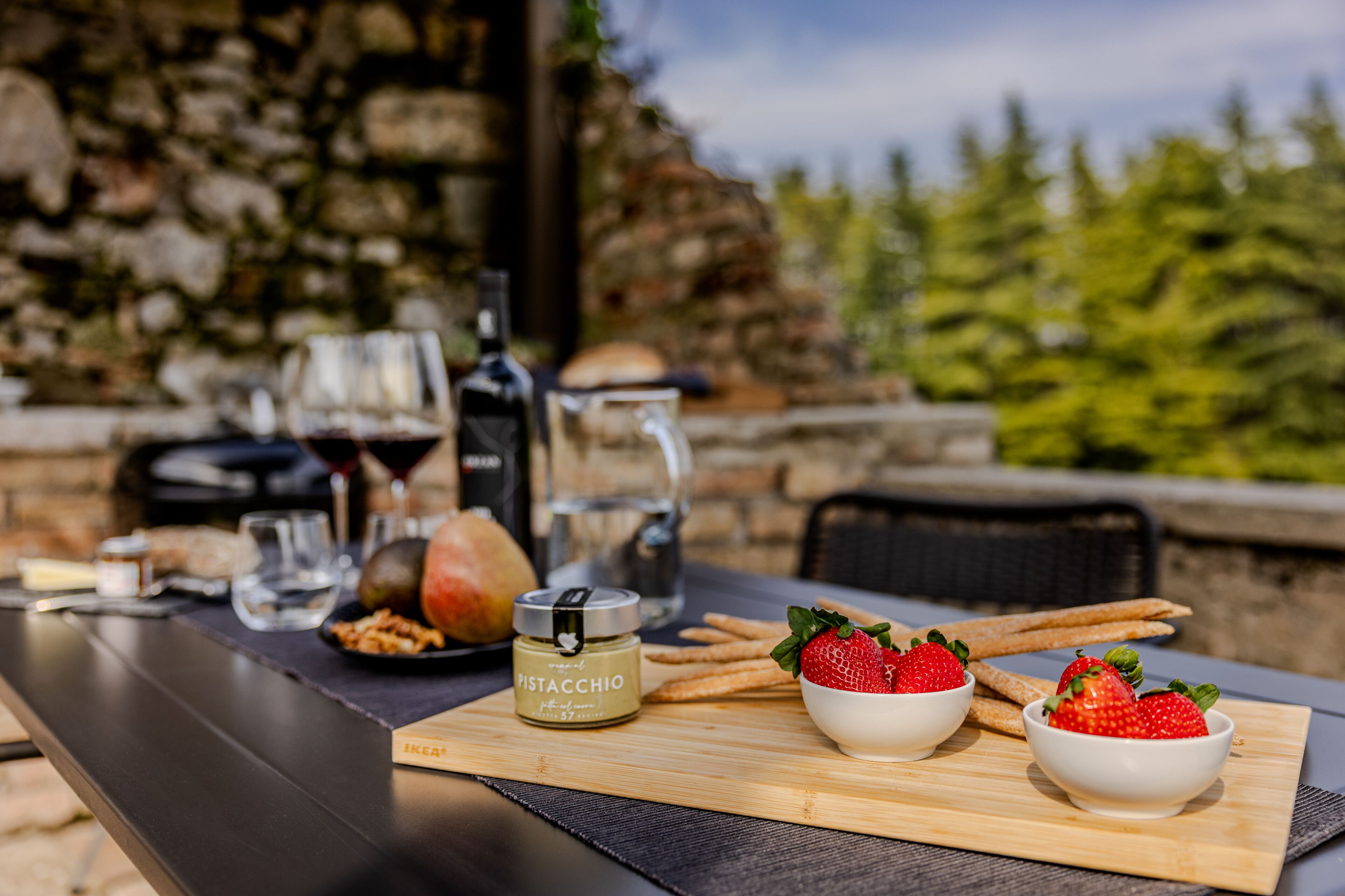 A table with pistachio spread, strawberries, crackers, and a bottle of wine, set against a stone wall and greenery.