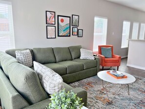 A modern living room with a sectional sofa, a small orange armchair, a coffee table, and a variety of framed artwork on the wall.