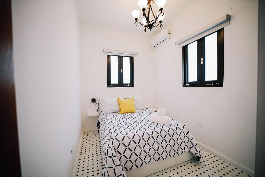 Una habitación pequeña y limpia con una cama, dos ventanas, una lámpara de techo y un radiador.