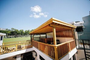 Una terraza de madera con barandilla, un área de estar techada y vistas a un campo verde.