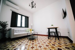 A living room with a sofa, a coffee table, a dining table with chairs, a potted plant, and a wall-mounted TV.